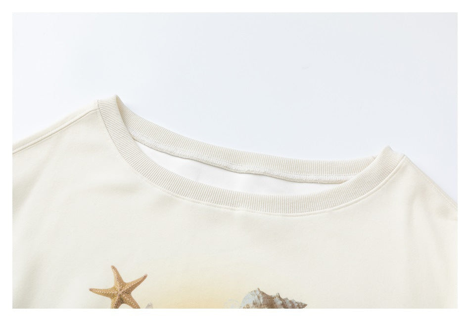 Close-up of a cream sweatshirt's ribbed crew neckline and the top of its starfish-and-shell print.