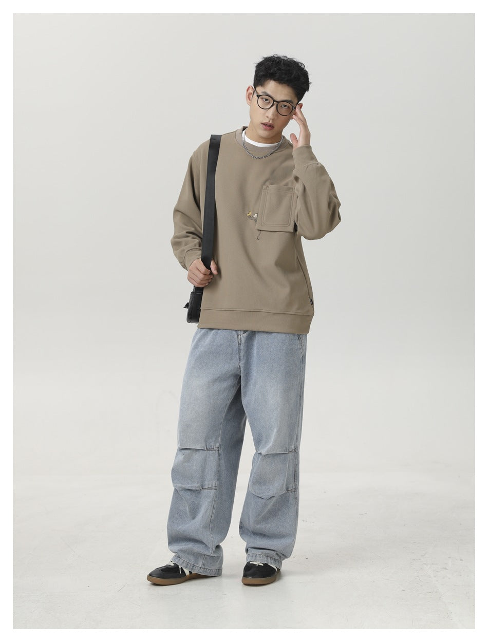 Full-length view of a man wearing a taupe pocket sweatshirt and loose light-wash jeans.