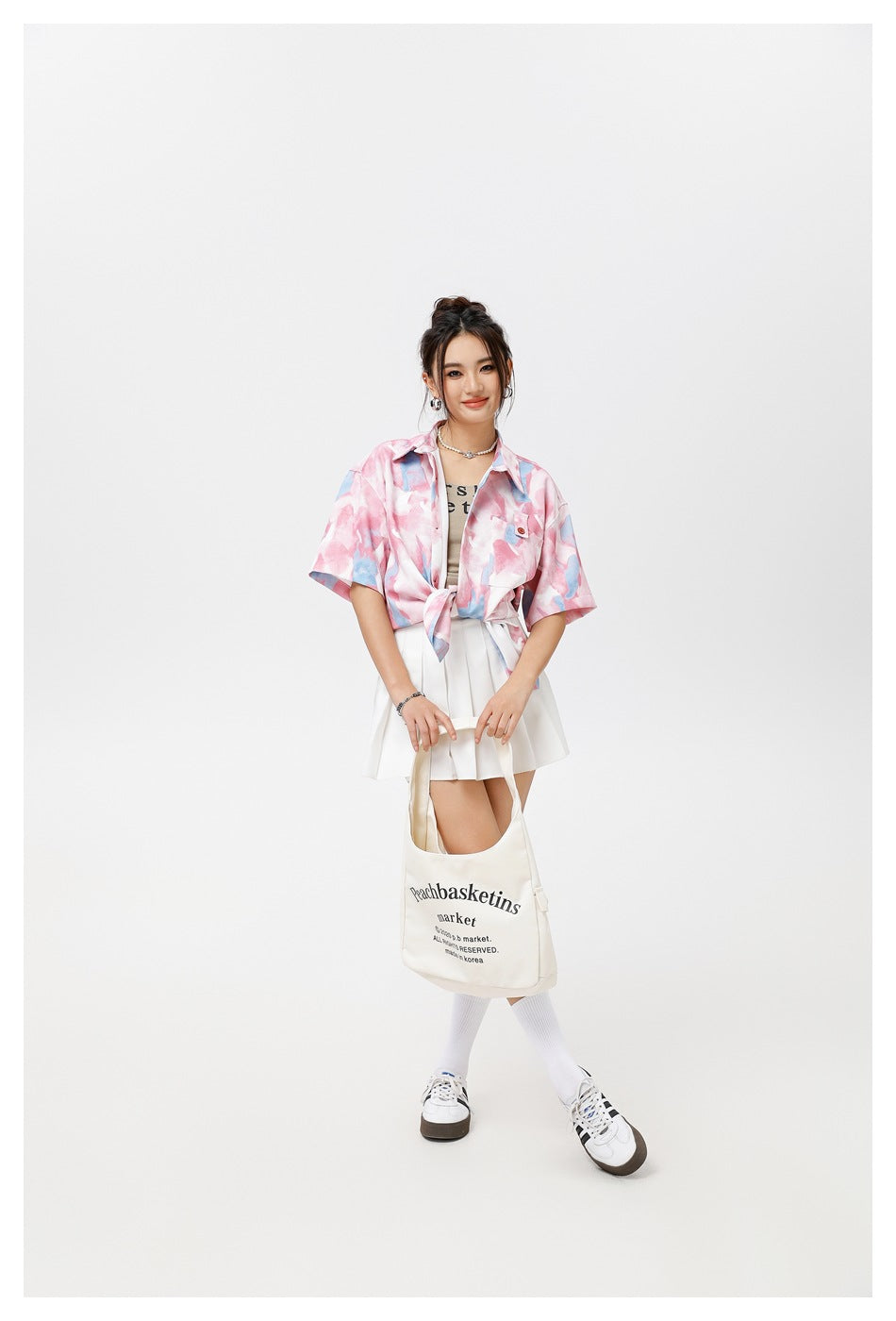 Model wearing a pink and blue abstract-print tied shirt with a white pleated skirt and carrying a cream tote.