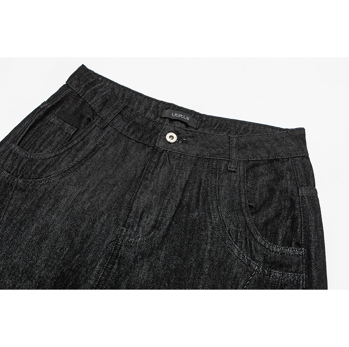 Close-up of black trousers showing the waistband, metal button, front pleats and pockets.