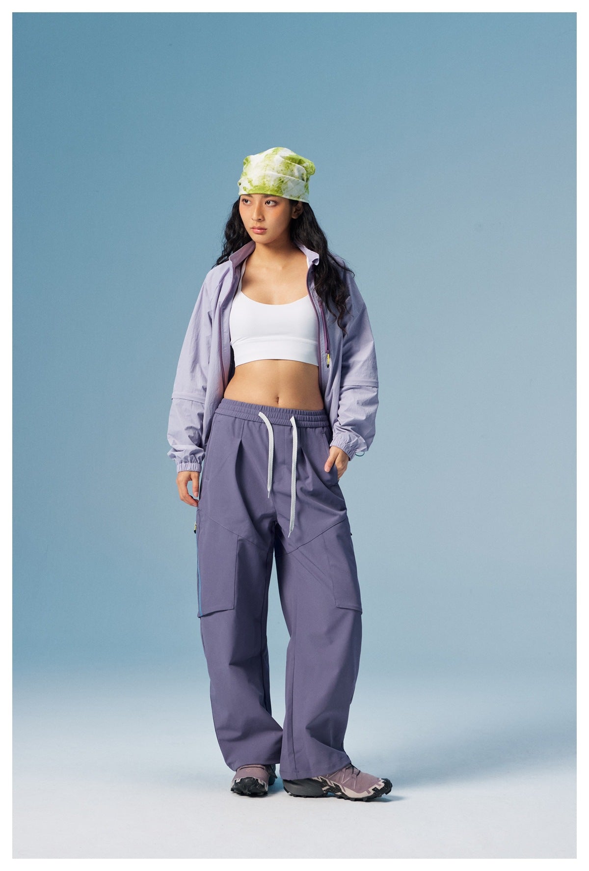 Model wearing a pale lavender zip jacket and loose purple cargo pants against a blue studio background.