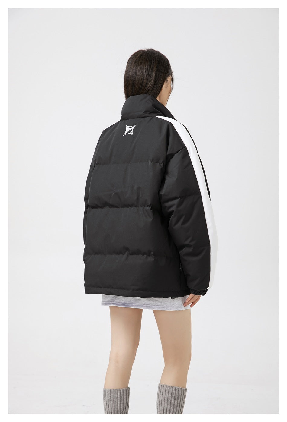 Closer back view of a model wearing a black padded jacket with white sleeve stripes and a small upper-back emblem.