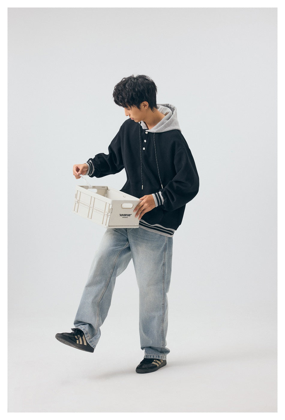 Model wearing a black and gray pullover hoodie with striped cuffs and hem, shown with a crate and light jeans.
