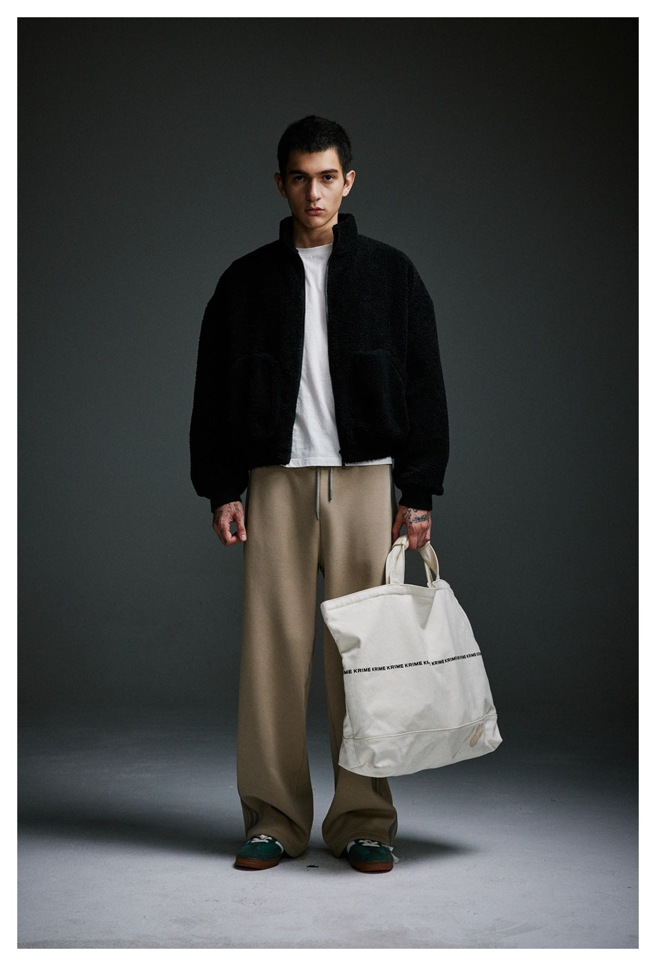 Model wearing the black fleece-like zip jacket open over a white tee with beige wide pants, holding a canvas tote