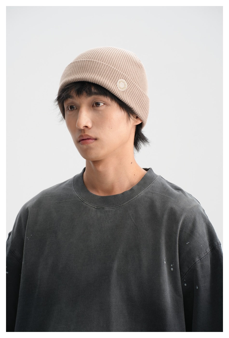 Close-up of a model wearing a charcoal T-shirt and ribbed beige beanie with a small circular patch.