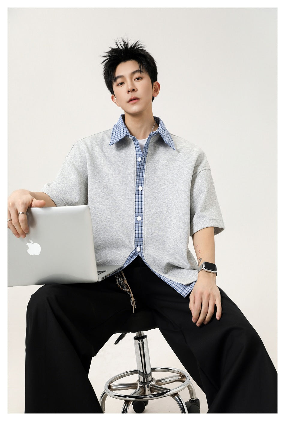 Man seated on a stool holding a laptop, wearing a gray short-sleeve top with blue plaid layers and black wide-leg pants