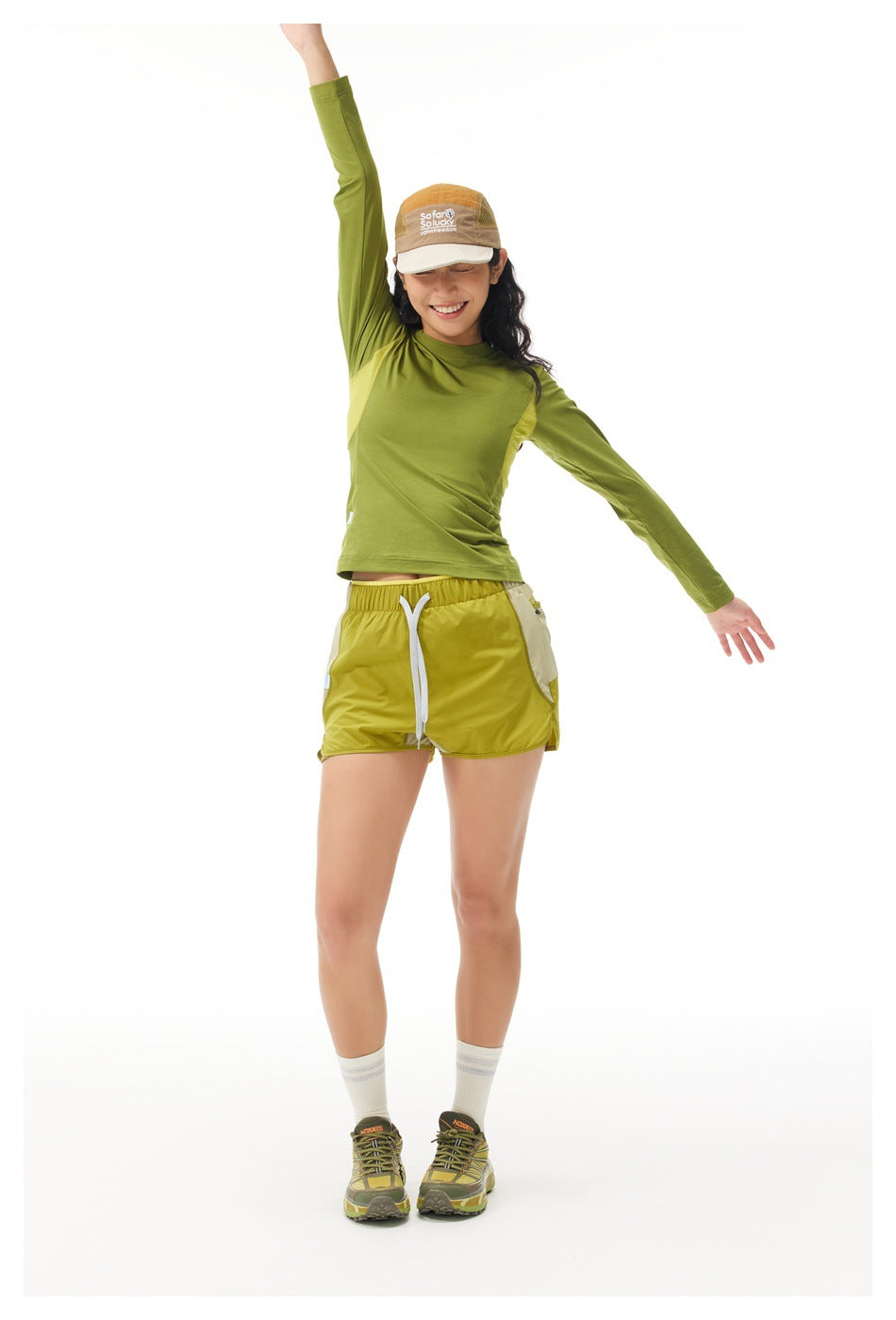 Front view of a model wearing an olive-green long-sleeve top and yellow-green drawstring shorts, posing with one arm raised.