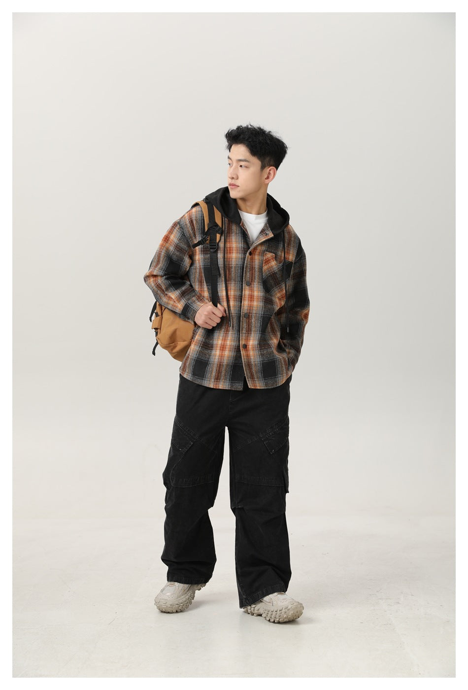 Full-length view of a man in a black, gray, and orange plaid hooded overshirt, black cargo pants, and tan backpack.