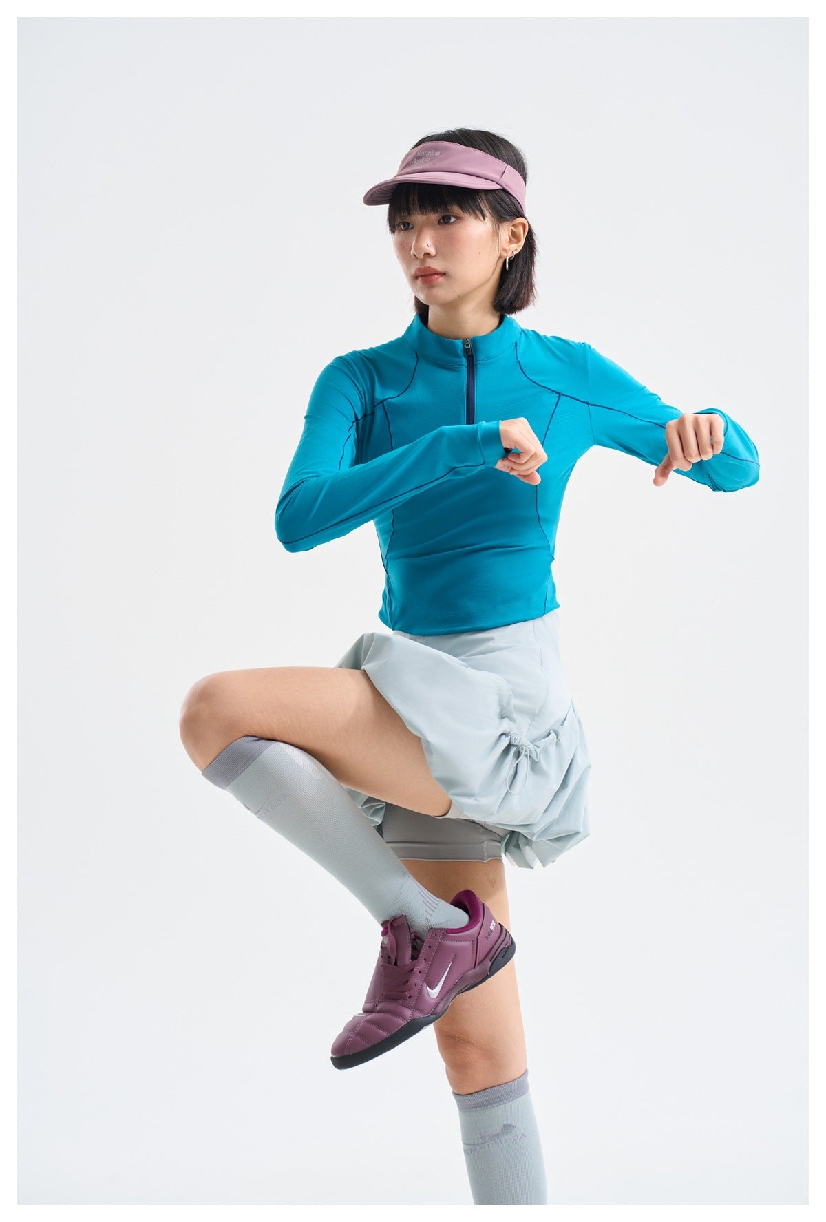 Model wearing a bright teal long-sleeve quarter-zip top with dark seam lines and a pale blue skirt.