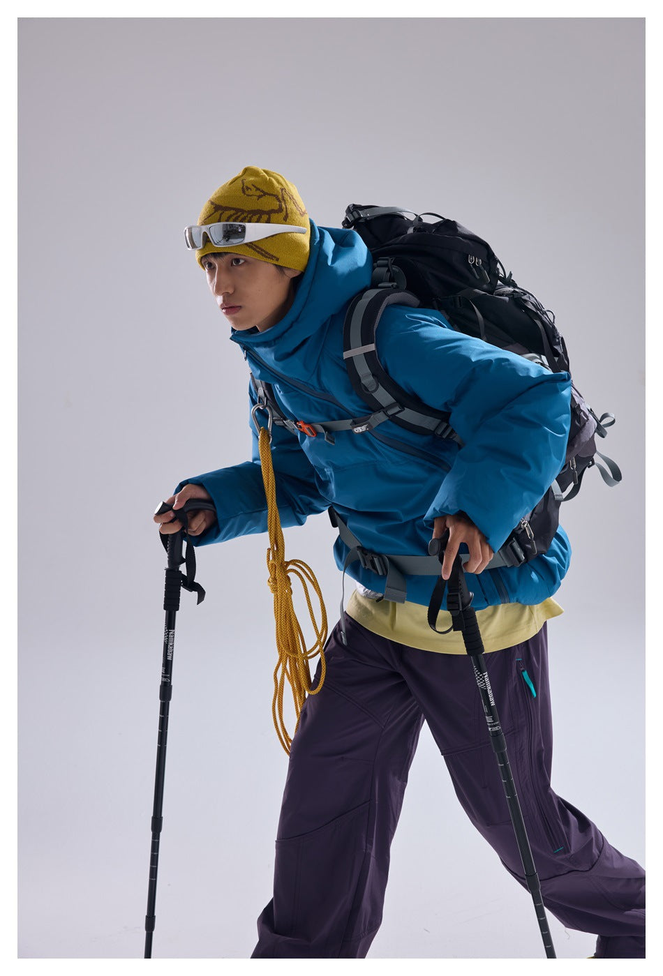 Model wearing a bright blue hooded jacket and purple pants, leaning forward with a backpack and trekking poles.