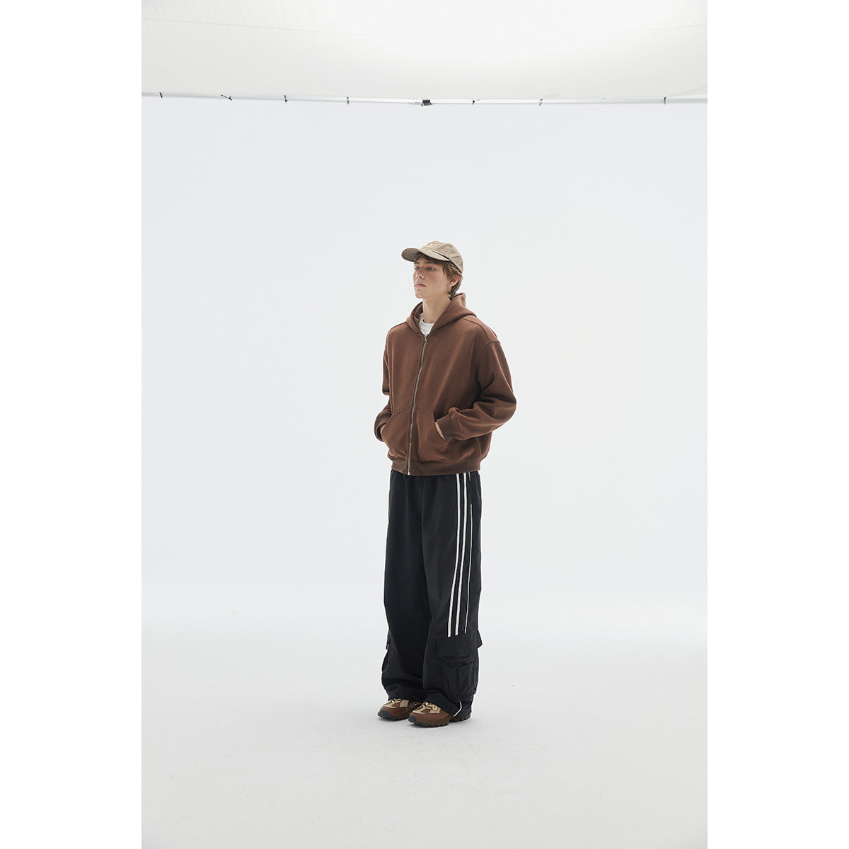 Model wearing a brown zip-up hoodie with a hood and front pockets, styled with black striped cargo pants.