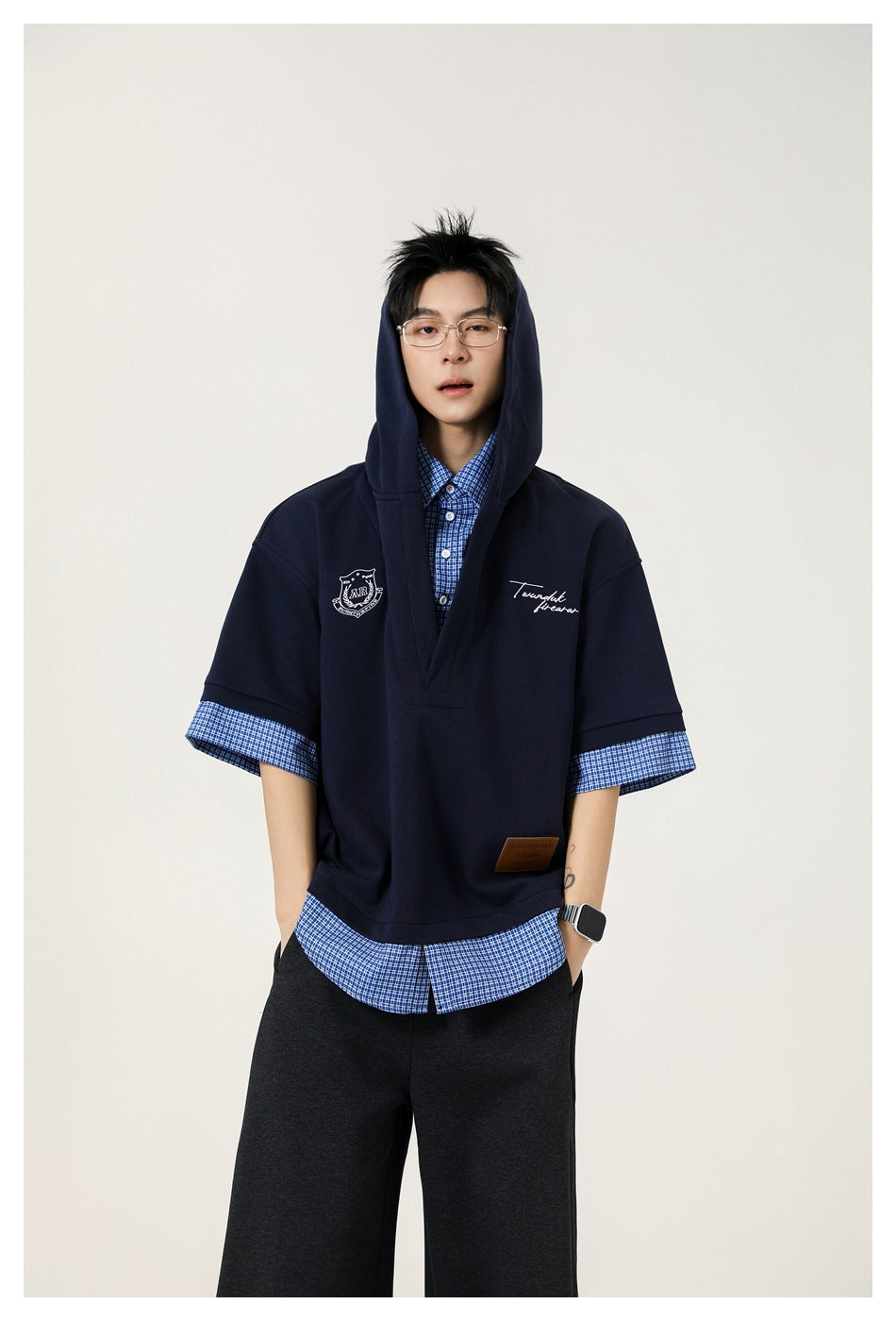 Model wearing navy layered hooded top with blue check shirt layer, hood up, with dark wide-leg pants