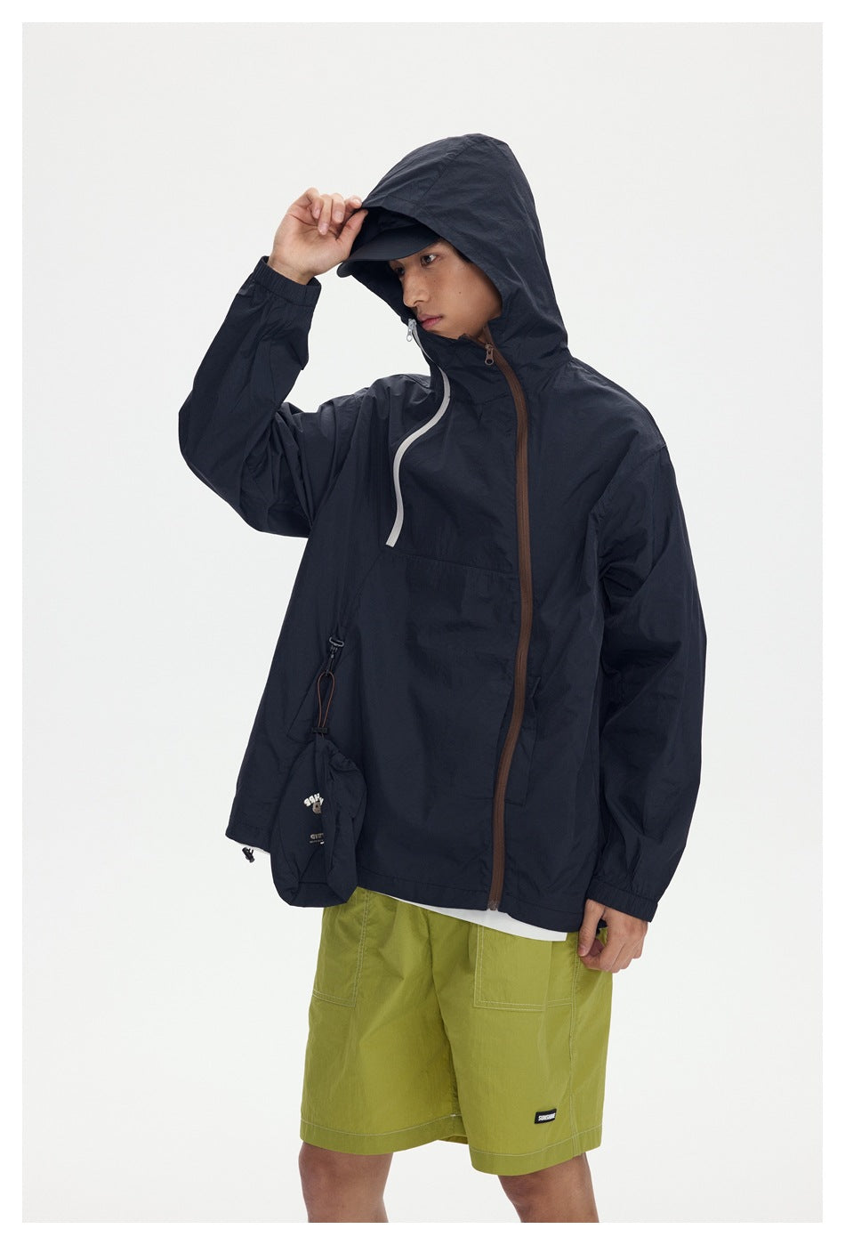 Model wearing a dark hooded jacket with contrasting zippers and lime-green shorts, adjusting his cap.