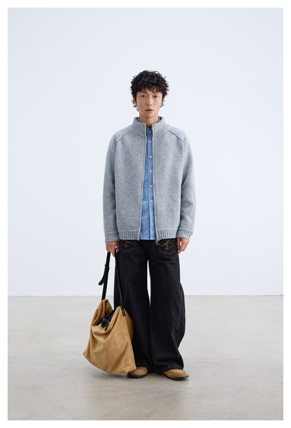 Full-length model wearing a light gray zip-front cardigan, blue shirt, and wide black jeans, with a tan bag.
