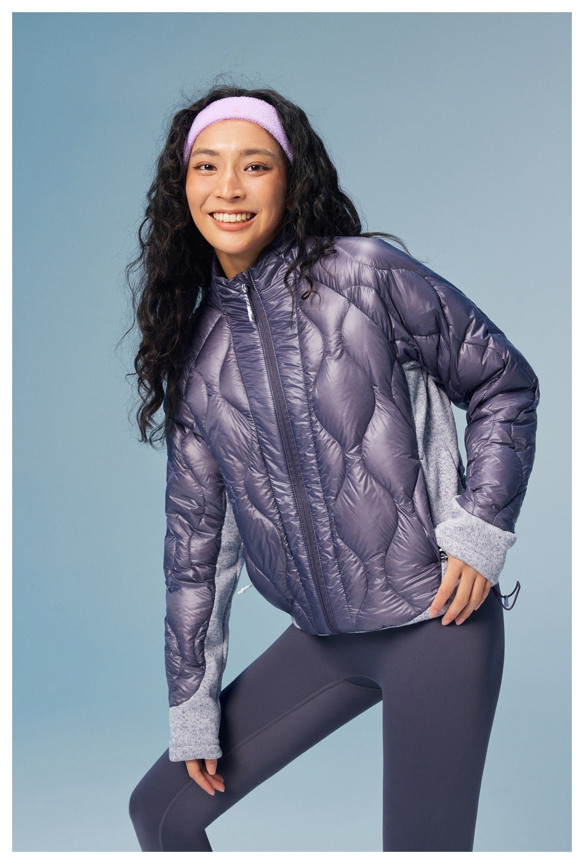 Model leans forward wearing a purple quilted jacket with gray panels and a front zipper.