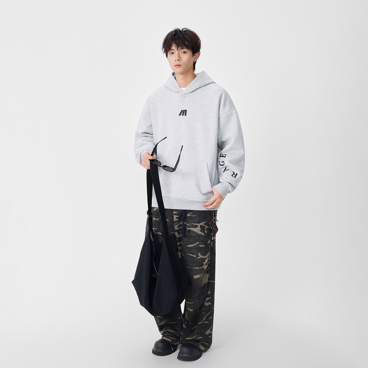 Model wearing a light gray oversized hoodie with a small chest emblem and sleeve lettering, paired with camouflage pants.