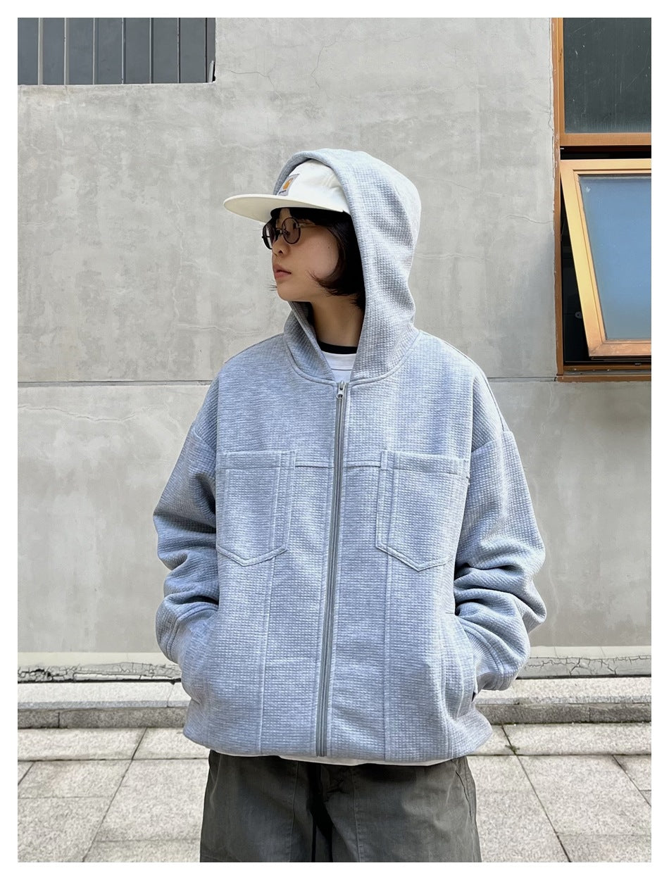 Person wearing gray waffle-knit zip-up hoodie with hood up, hands in side pockets, front view