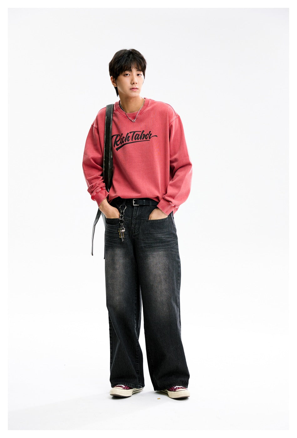 Model wearing red crew-neck sweatshirt with black script print, wide-leg dark jeans and sneakers, full-length view