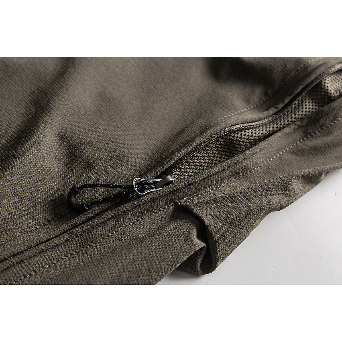 Close-up of an olive-gray trouser pocket with a zipper, mesh lining, and a pull cord.