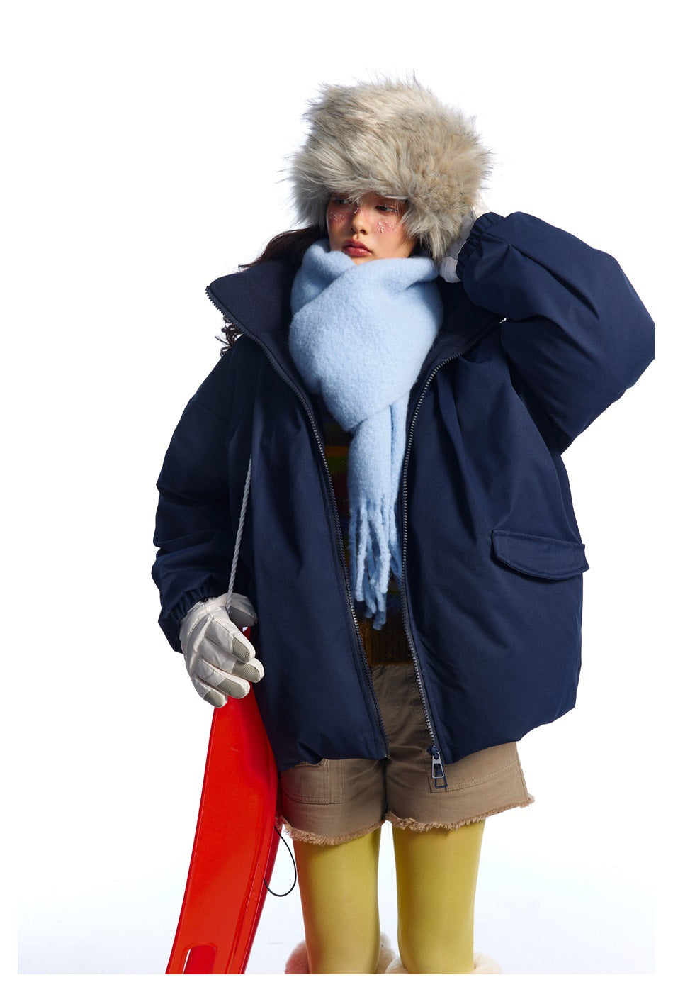 Model wearing an open navy jacket and pale blue scarf, with a fur hat, gloves and beige shorts.