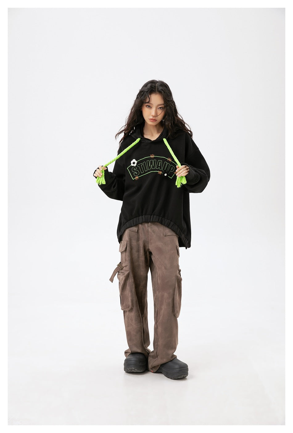 Model wearing a black hoodie with neon green drawstrings held out, paired with brown cargo pants