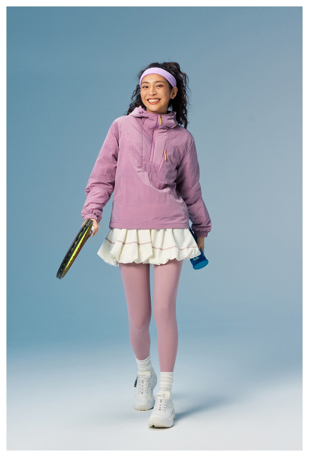 Model wearing a lilac hooded pullover, white skirt, and lilac leggings while holding a tennis racket.