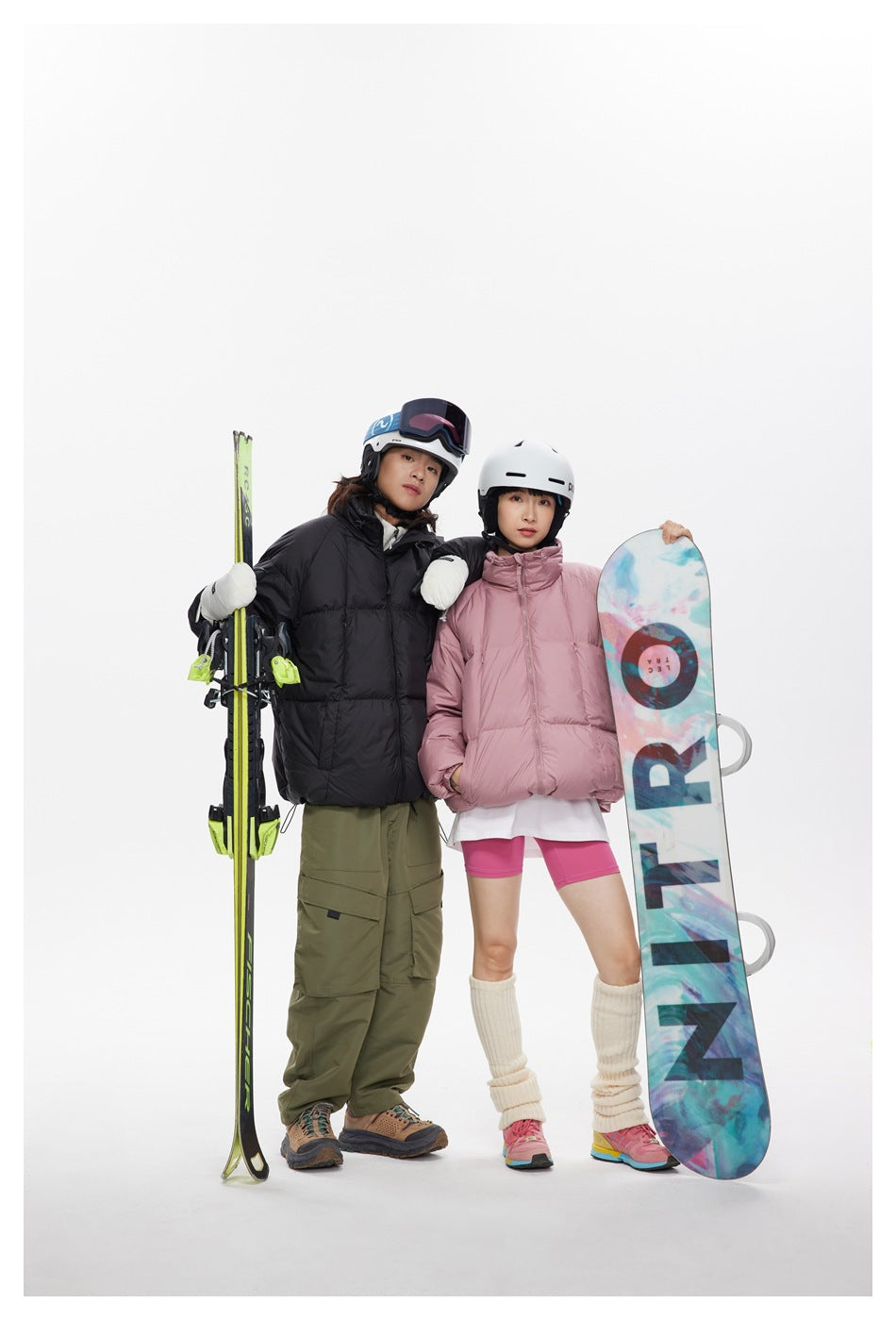 Two models in black and dusty pink puffer jackets pose with skis and a colorful snowboard.
