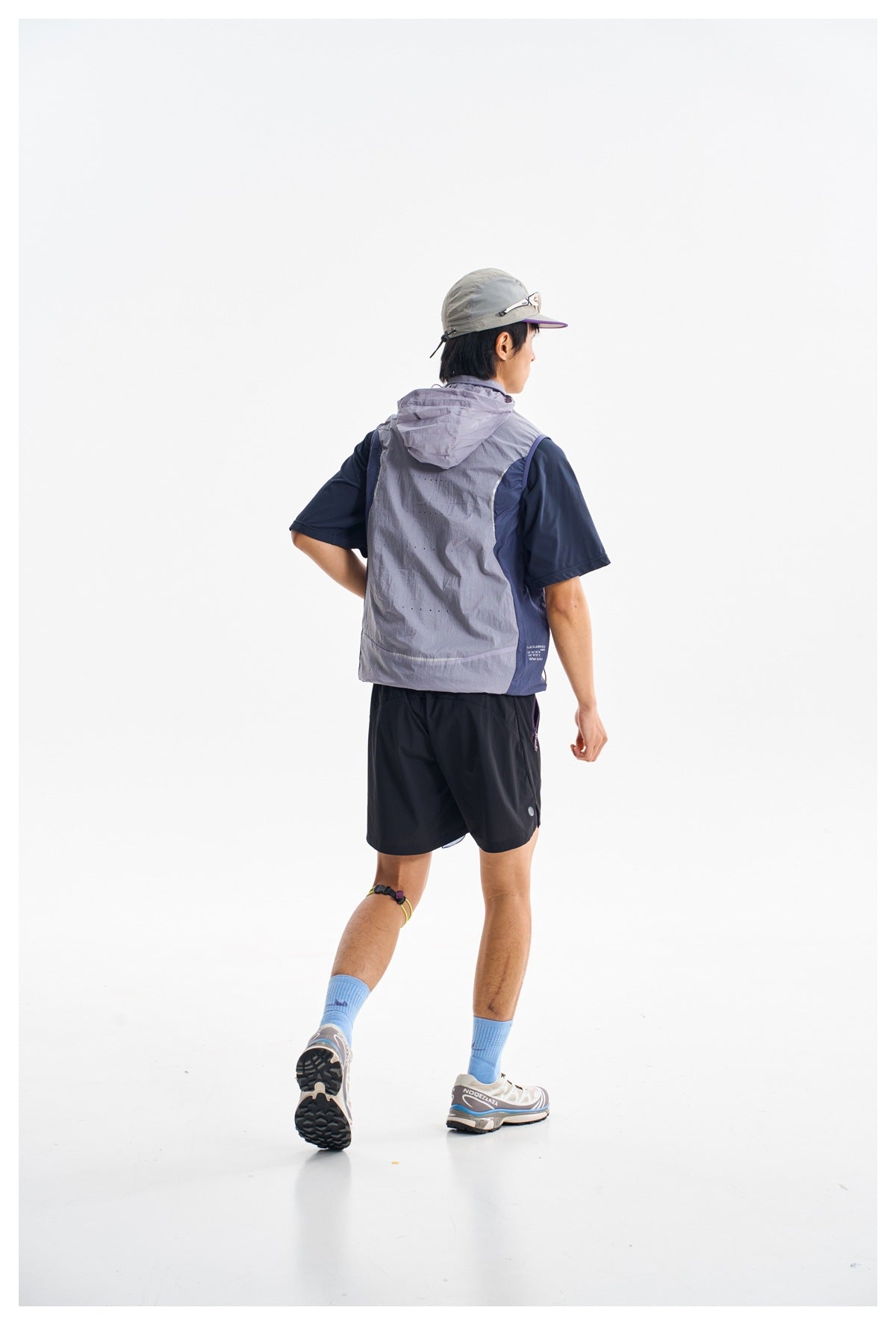 Rear view of a man in a light gray hooded vest over a navy shirt, navy shorts, blue socks and a gray cap.