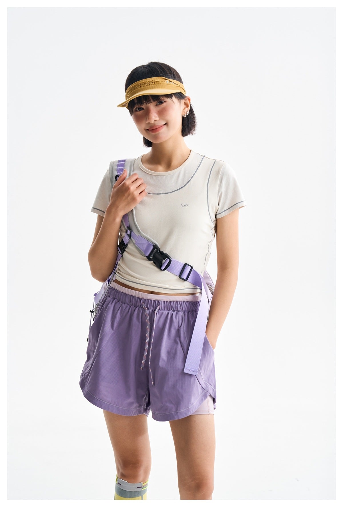 Closer view of a model in a cream athletic T-shirt and purple shorts, wearing a lilac crossbody harness and yellow visor.