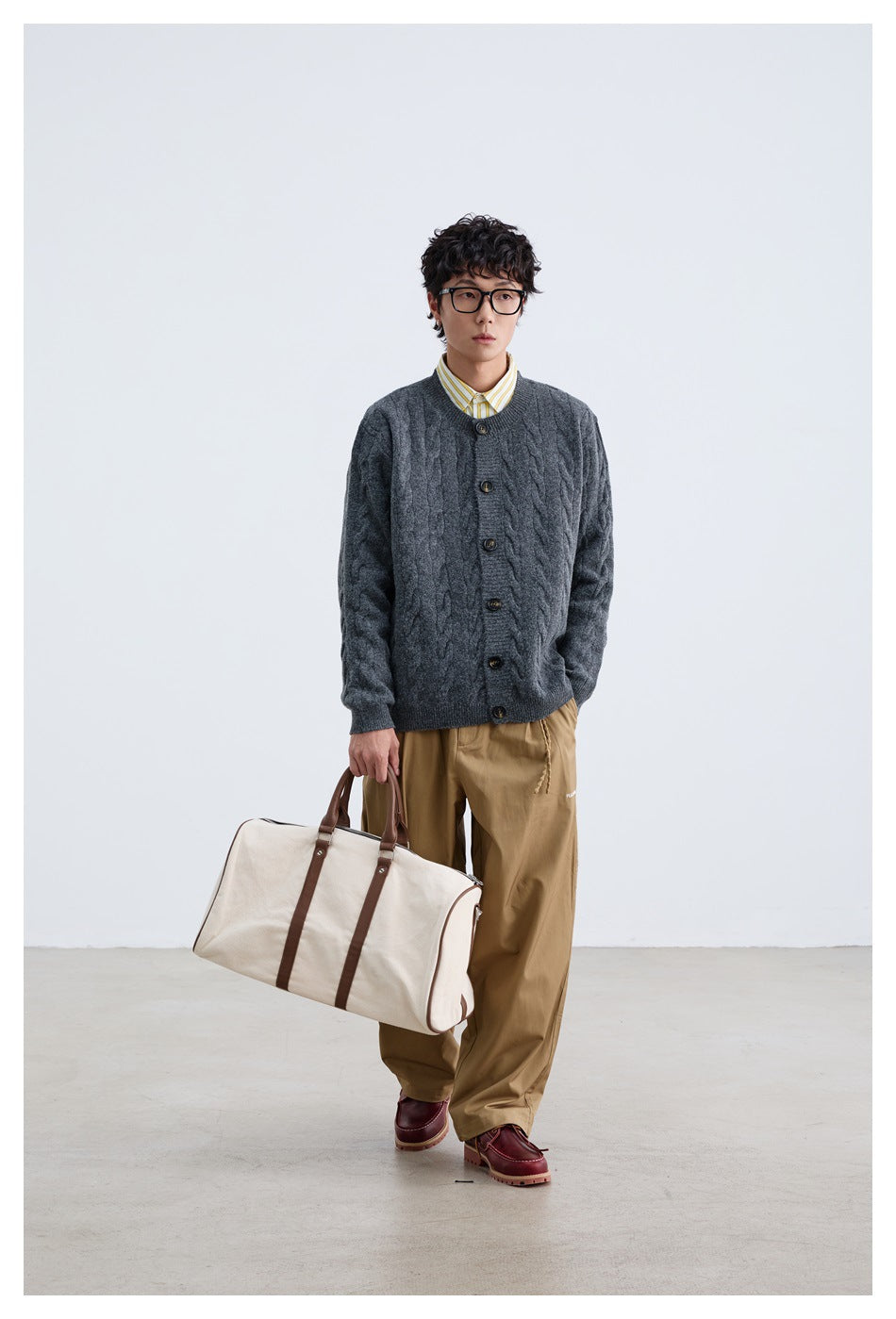 Man wearing dark gray cable-knit button cardigan over a striped shirt with tan trousers, holding a canvas duffel bag