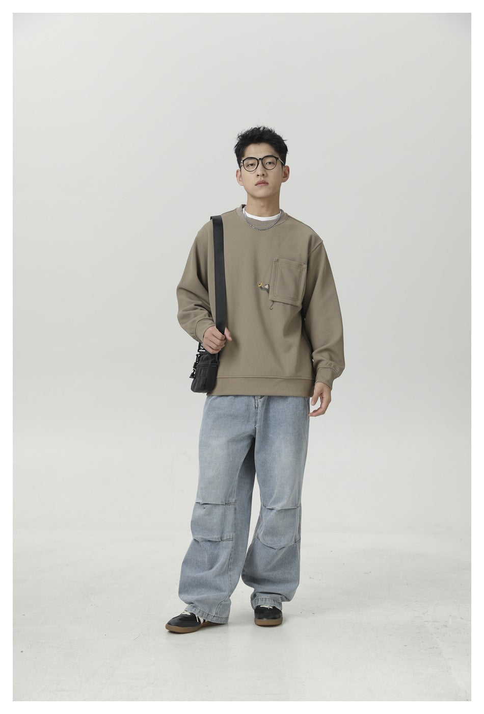 Full-length view of a man wearing a taupe pocket sweatshirt, loose light blue jeans, glasses, and a black shoulder bag.