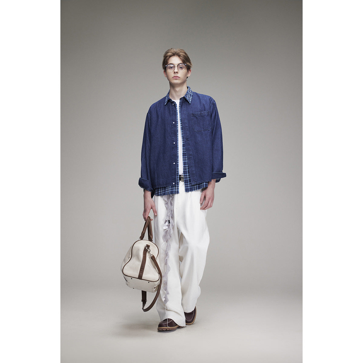 Full-length model in a navy denim overshirt layered over a blue plaid shirt and loose white trousers, carrying a cream bag.