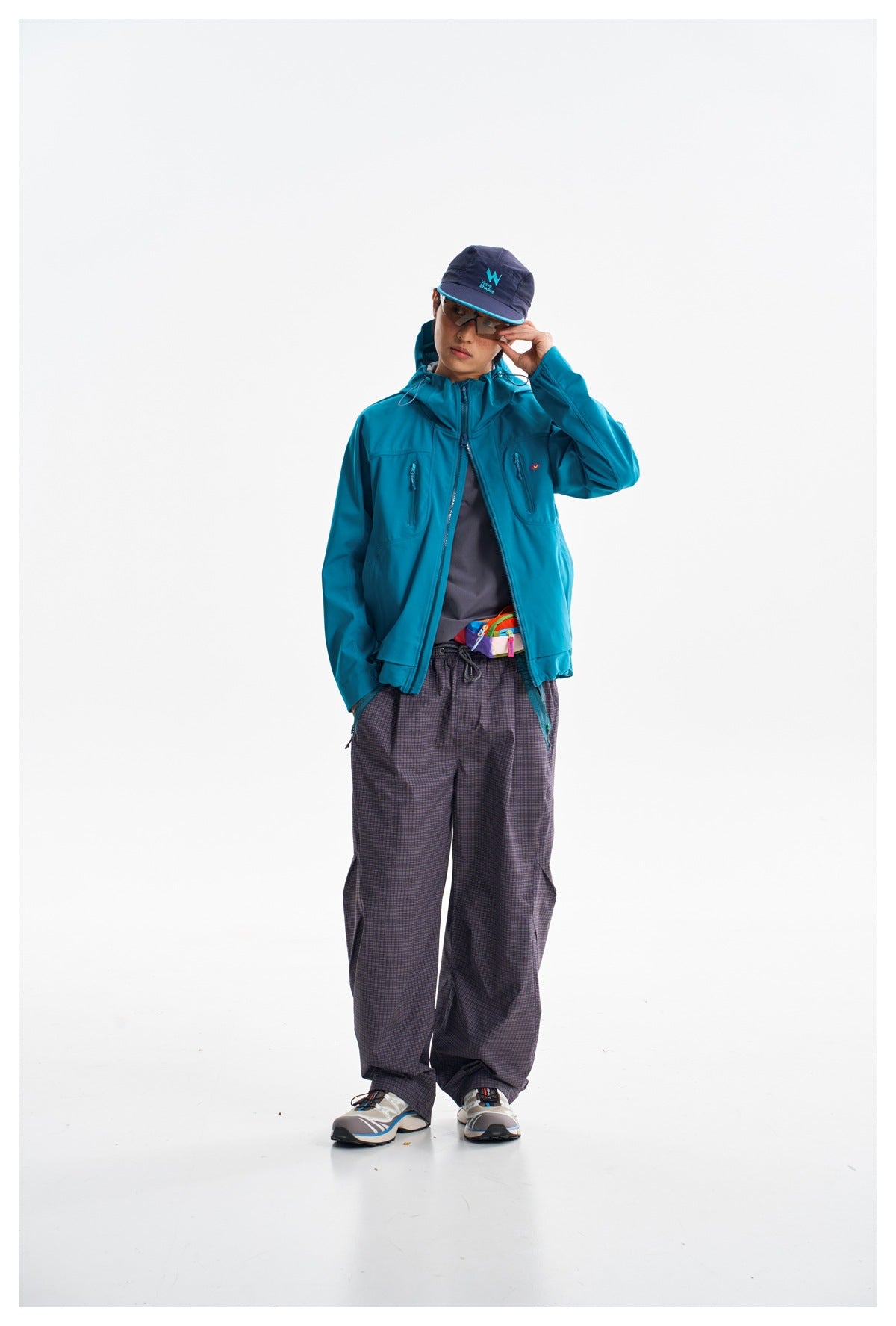 Front view of a model wearing a teal hooded zip jacket with chest pockets and plaid trousers.