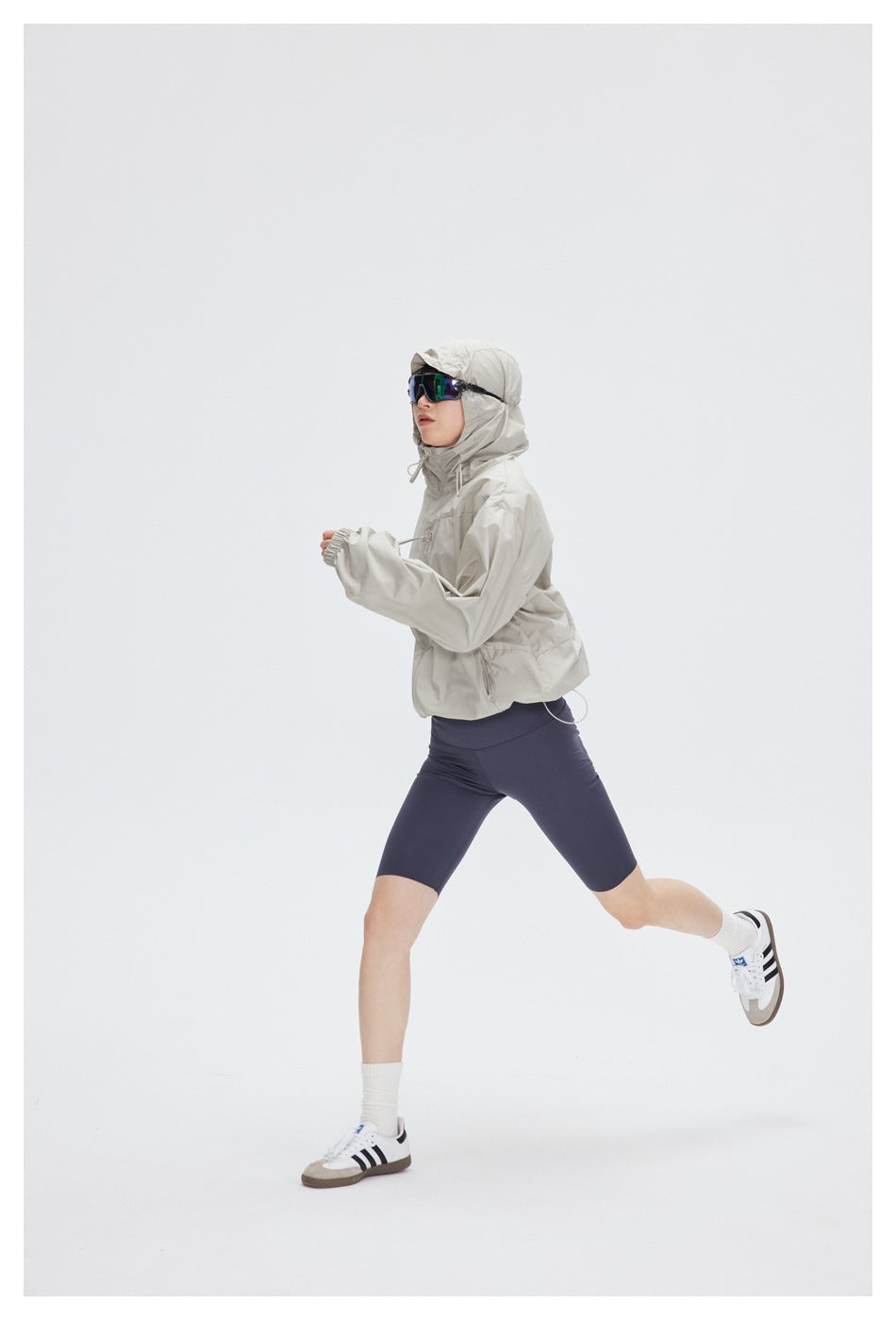 Model in a light beige hooded windbreaker and navy cycling shorts, shown mid-stride.