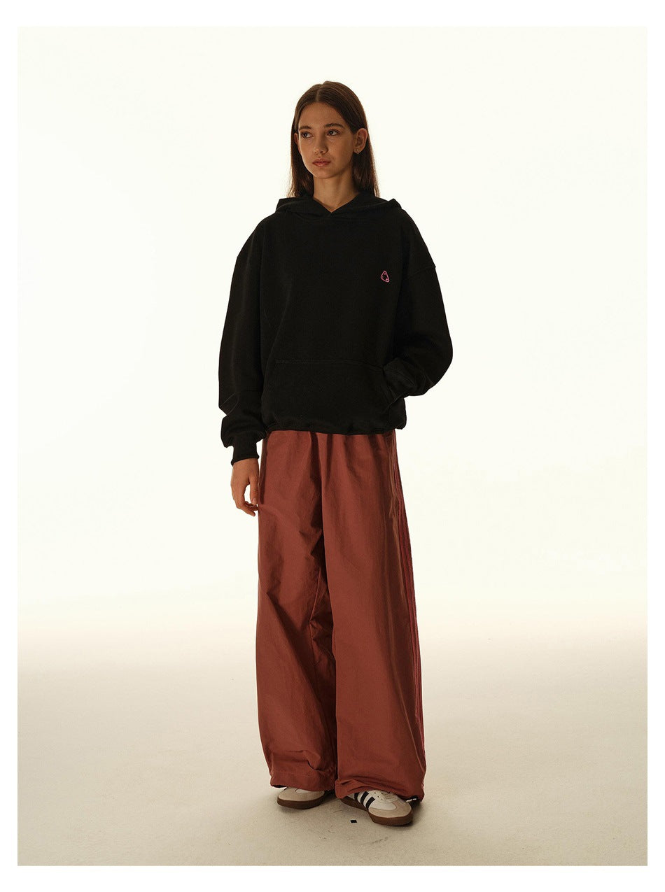 Model wearing a black oversized hoodie with a small chest graphic and wide rust-red trousers, front view.
