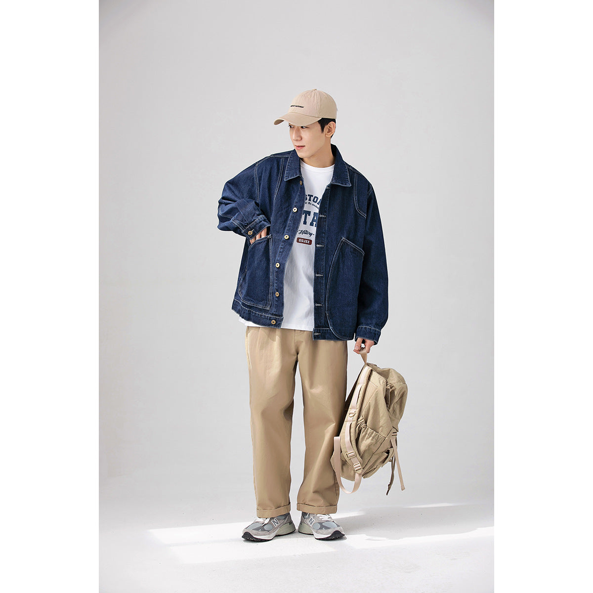 Full-length view of a model in a dark indigo denim jacket, beige trousers, and beige cap, holding a backpack.