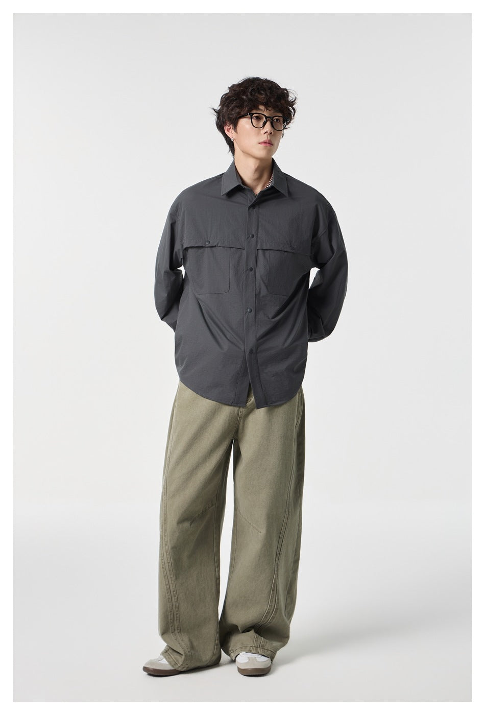 Model wearing dark gray oversized button-up shirt with chest pockets and wide olive pants, hands behind back