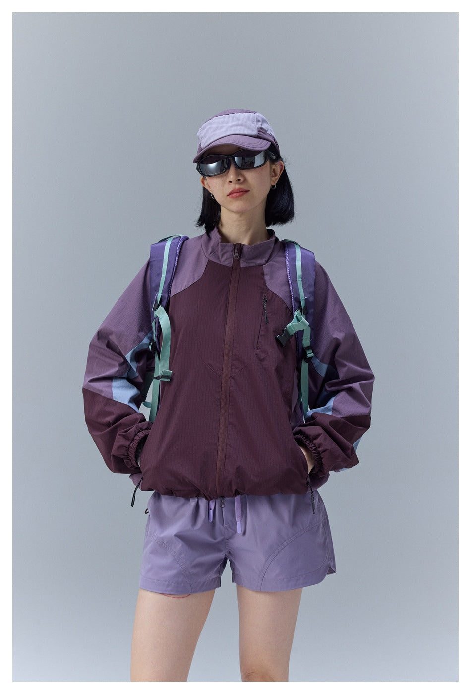 Front view of a model wearing a plum and purple color-block zip jacket with lavender shorts and a cap.