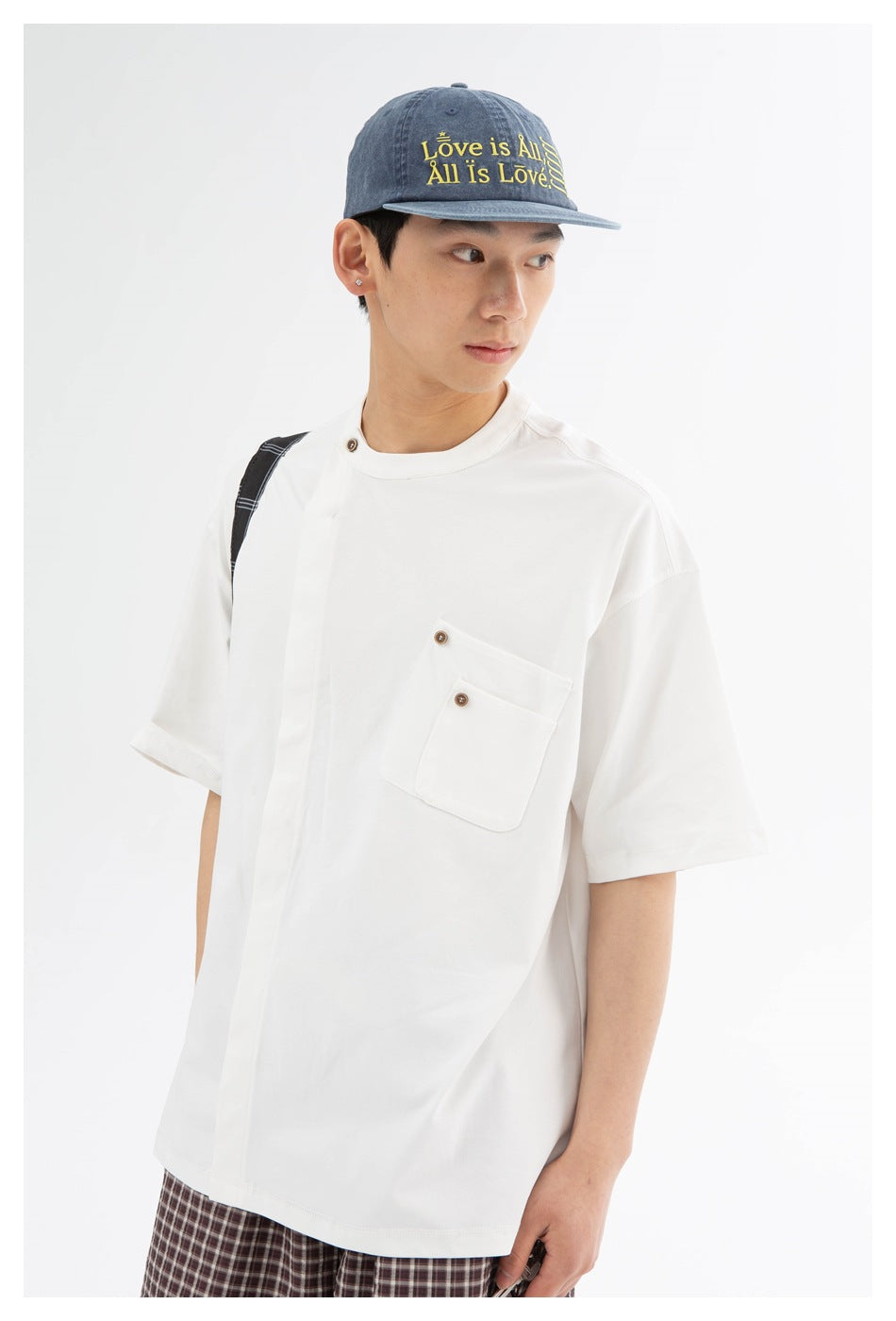 Model wearing an oversized white short-sleeve T-shirt with a button-detailed chest pocket; blue cap and plaid trousers.