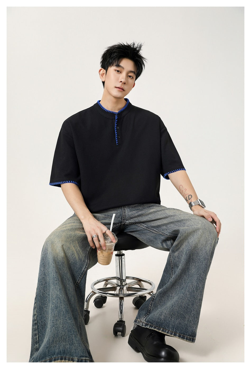 Seated model wearing a black short-sleeve henley with bright blue contrast stitching and wide-leg jeans.