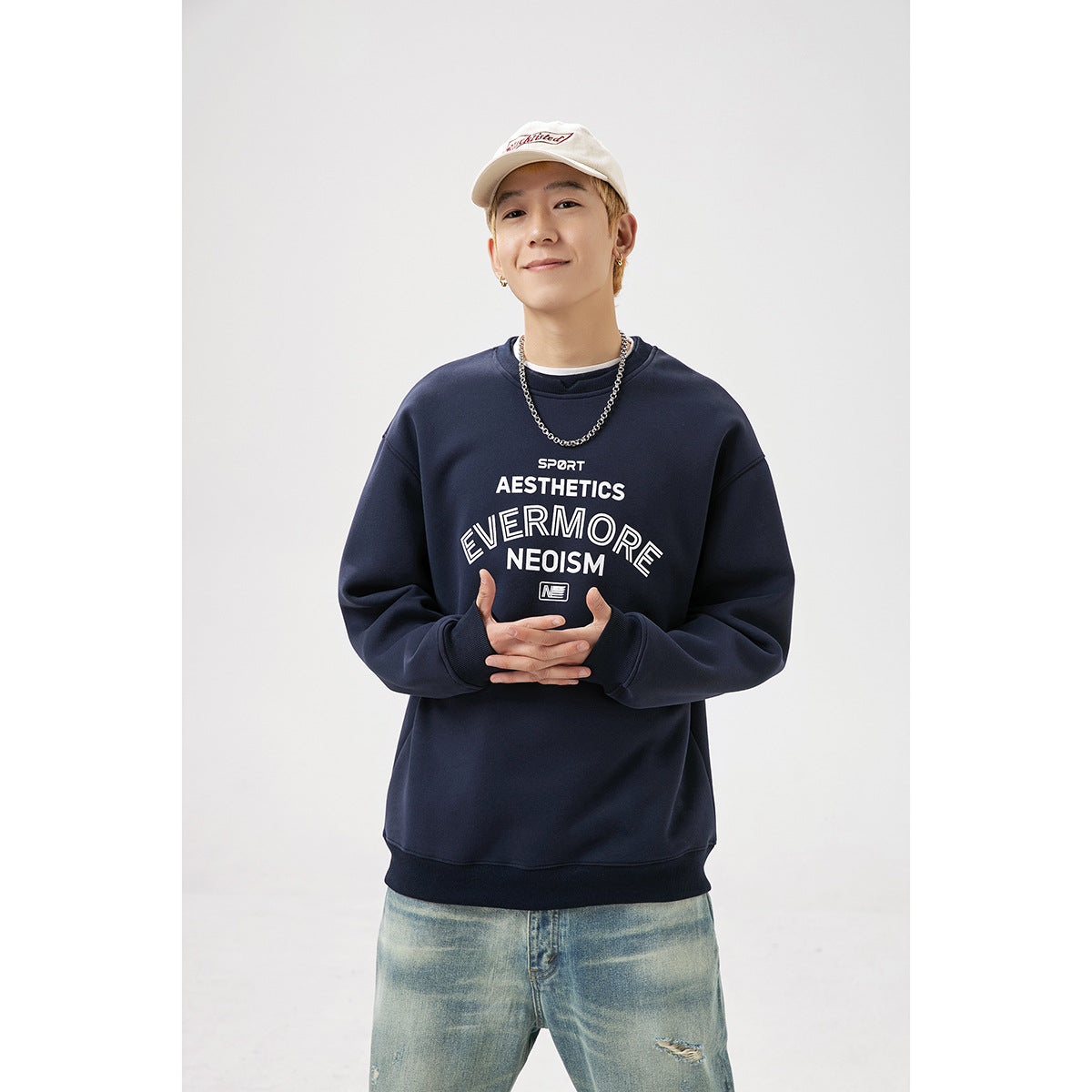 Model wearing a navy crewneck sweatshirt with white chest lettering, light jeans, and a cream cap.