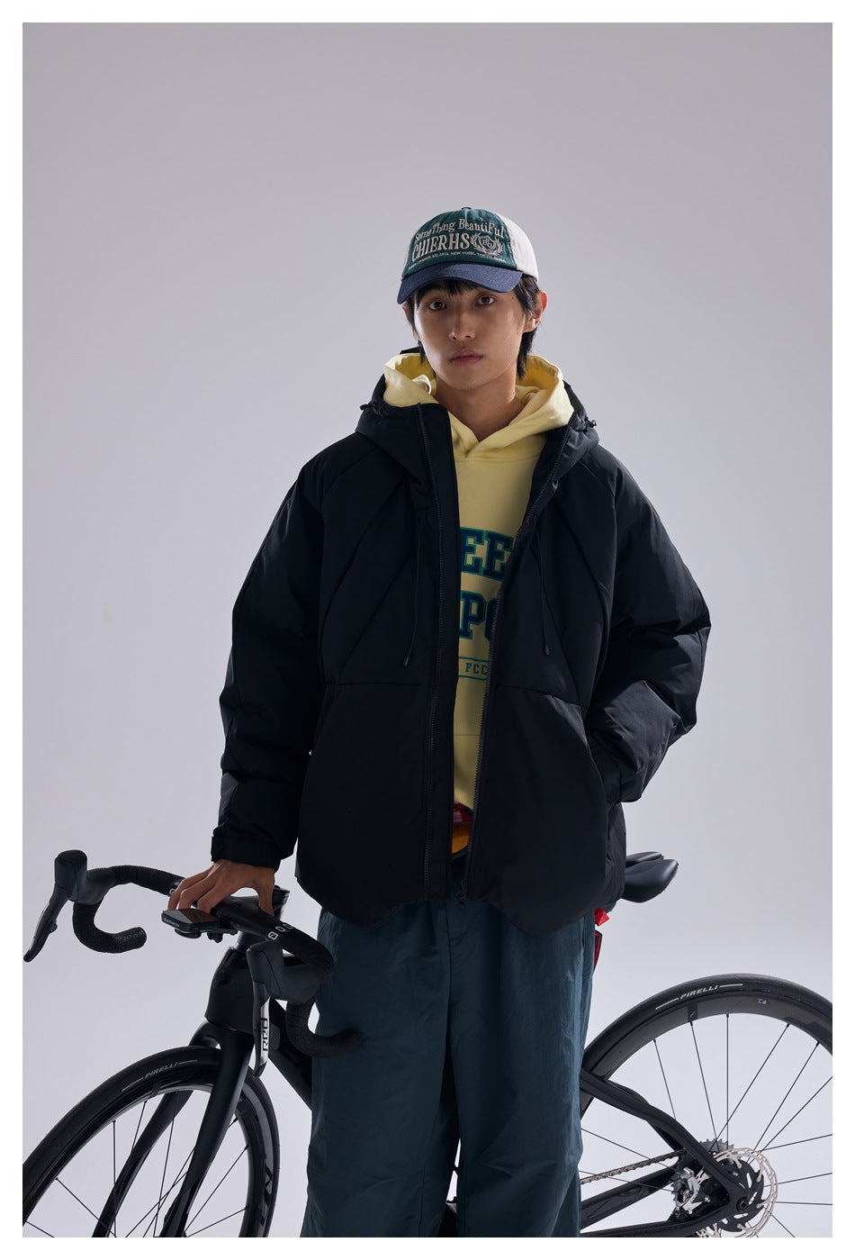 Model wearing an open black hooded puffer jacket over a pale yellow hoodie beside a bicycle.