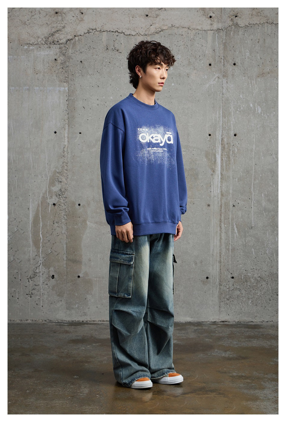 Model in a blue oversized crewneck sweatshirt with a white distressed front graphic, shown at a three-quarter angle.