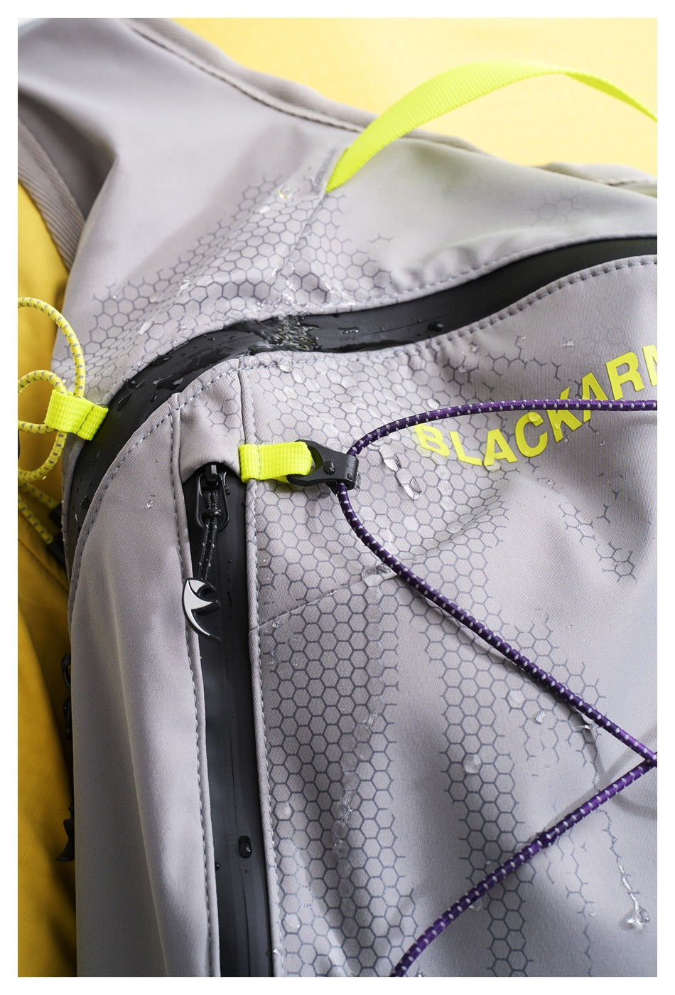 Close-up of a gray backpack showing water droplets, a black waterproof zipper, purple cord, and yellow accents.