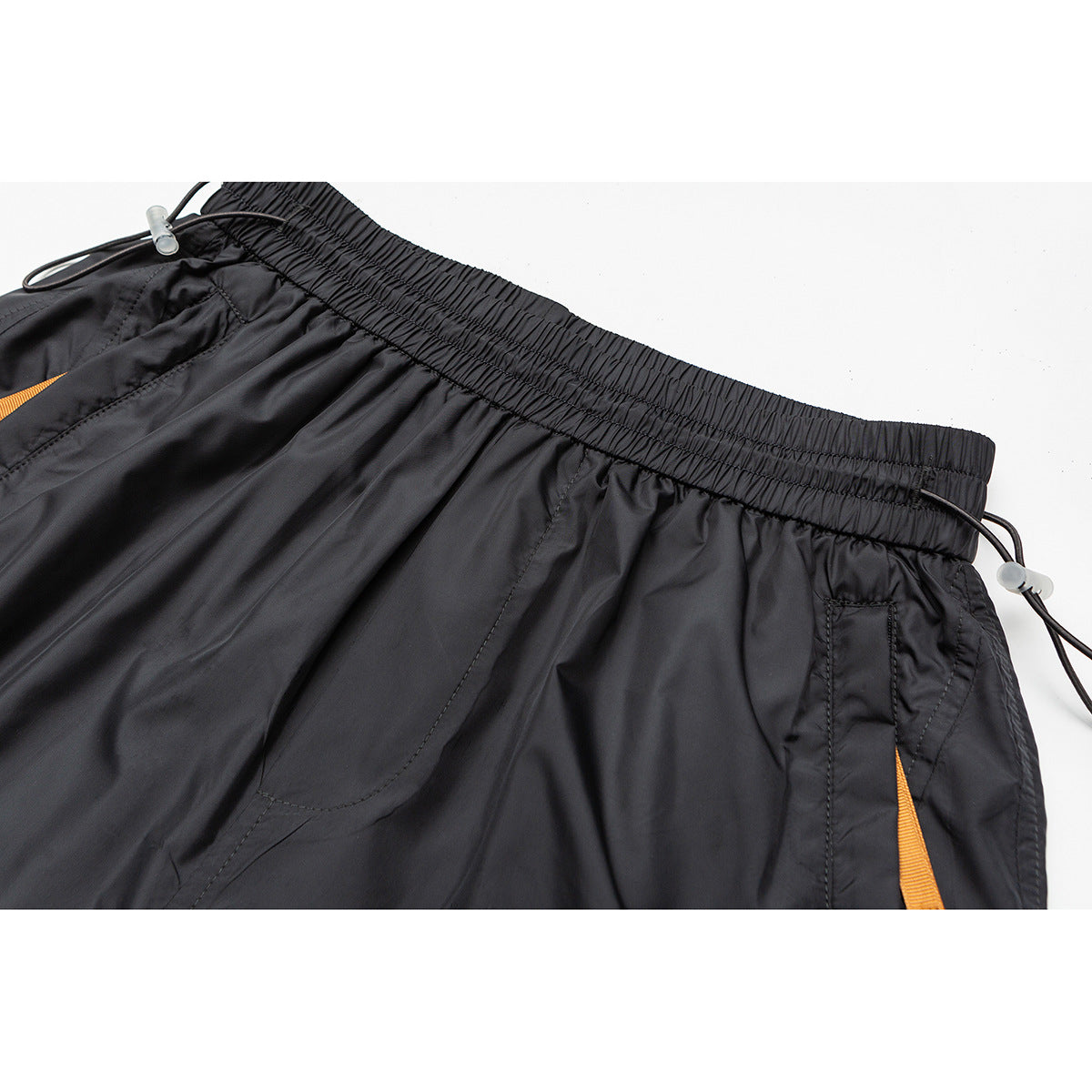 Close-up of black shorts showing the gathered elastic waistband, drawcords, pocket stitching, and orange trim.