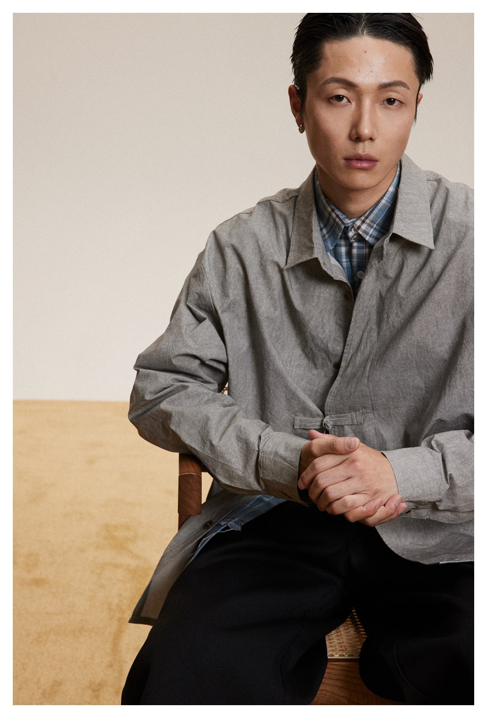Close view of a seated model wearing a light gray button-front jacket with a waist tab over a blue plaid shirt.