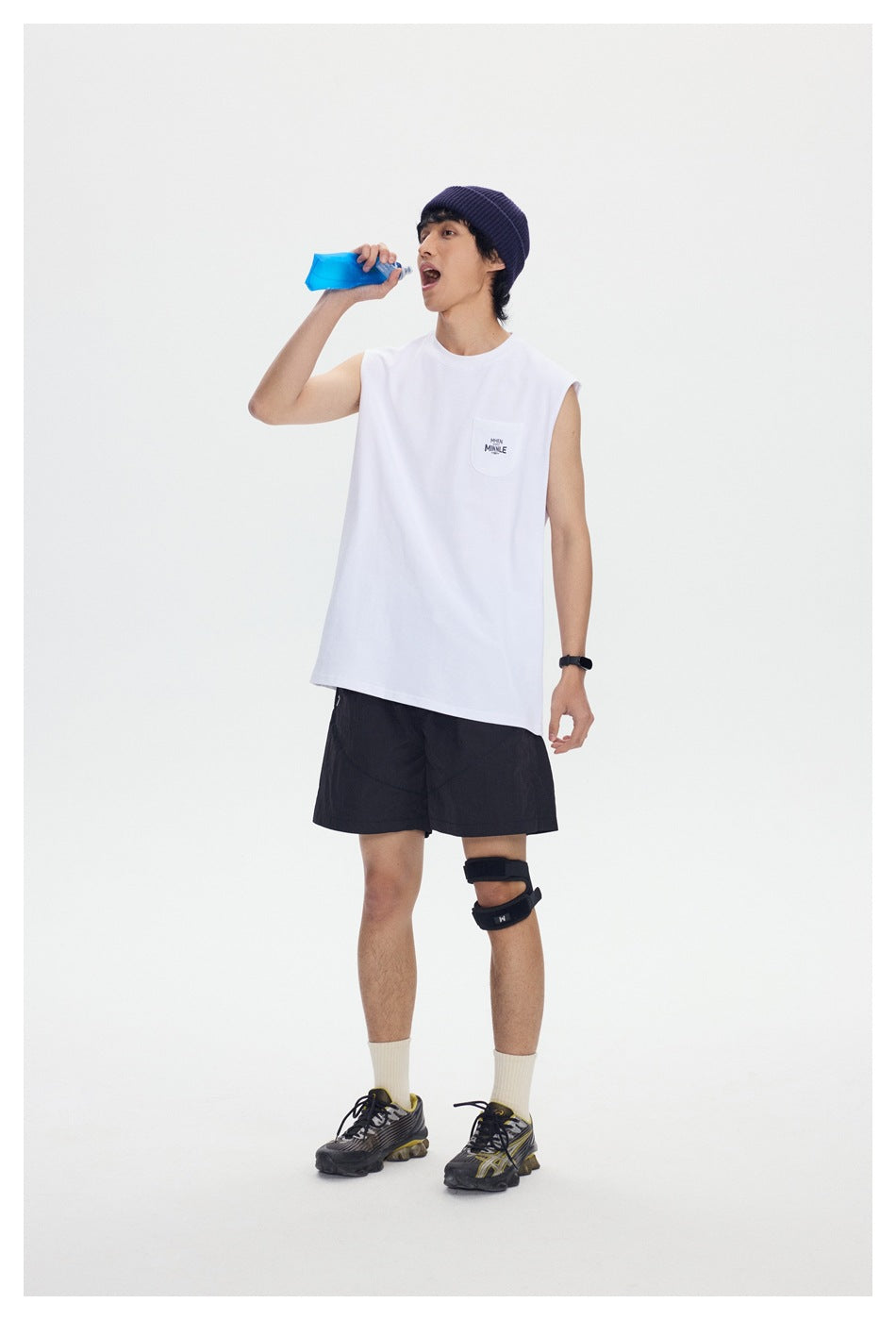 Model wearing a white sleeveless pocket shirt and black shorts, drinking from a blue bottle.