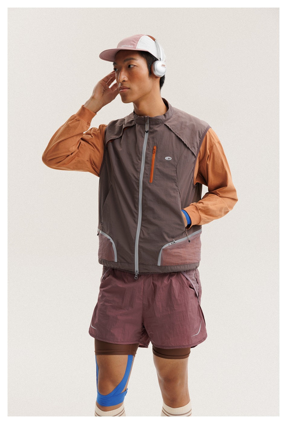 Close front view of a man wearing a taupe zip-front vest with orange sleeves and mauve shorts.