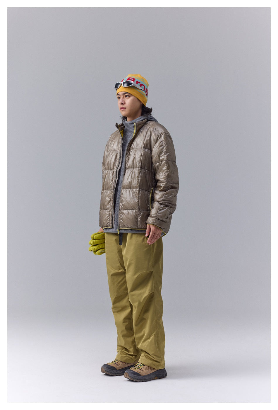 Full-length model wearing a taupe quilted jacket, gray top, olive pants, yellow beanie, and hiking shoes.
