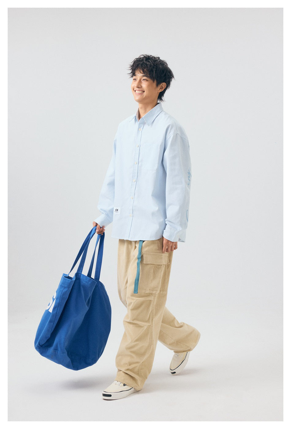 Smiling model wearing a pale blue button-up shirt and beige cargo pants, carrying a large blue tote.
